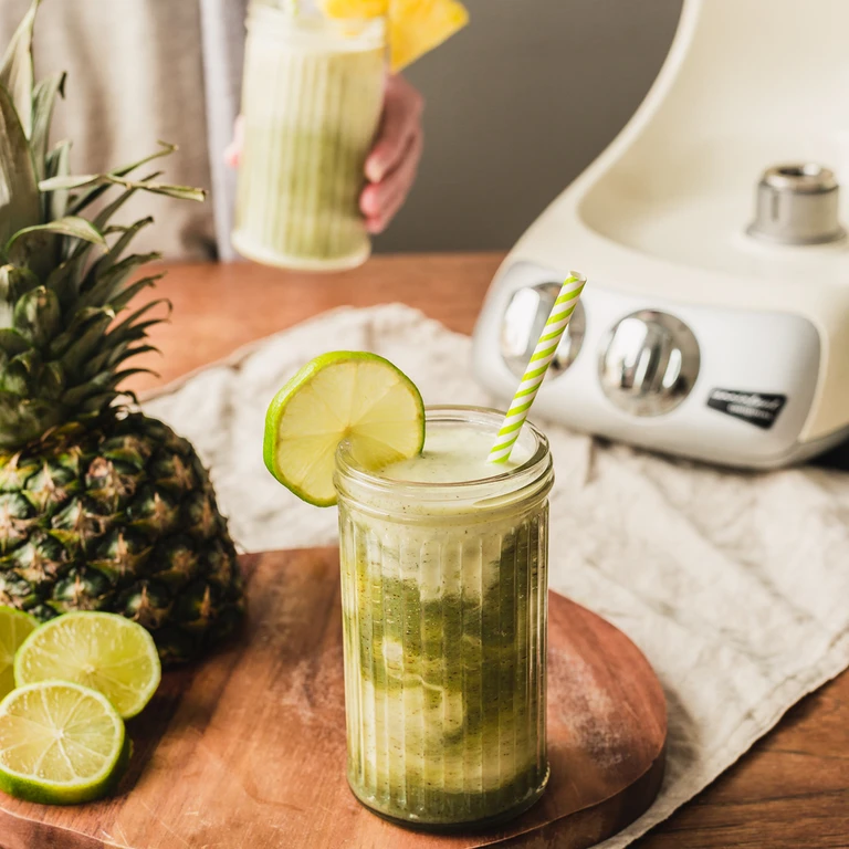 Green pineapple lime smoothie in a ribbed jar with a striped straw, made with an Ankarsrum stand mixer nearby.