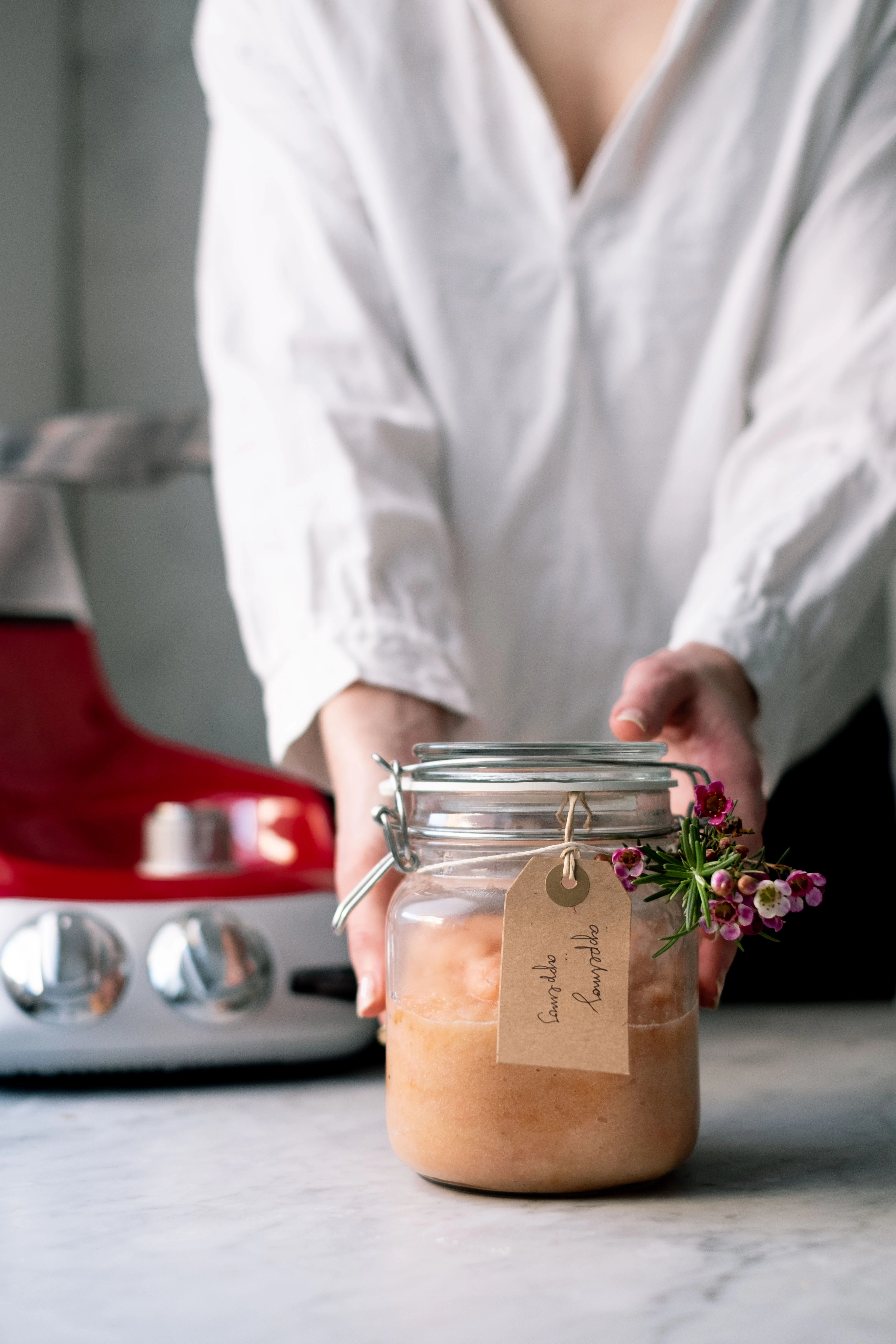 Woman presenting a labeled applesauce jar with herbs beside a red Ankarsrum stand mixer on a marble countertop.