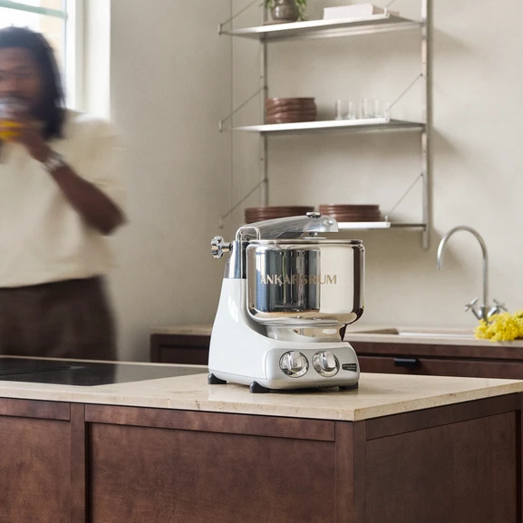 Ankarsrum Assistent Original in Glossy White on a light kitchen countertop in a kitchen with dark wood, with a person partially visible beside the machine.
