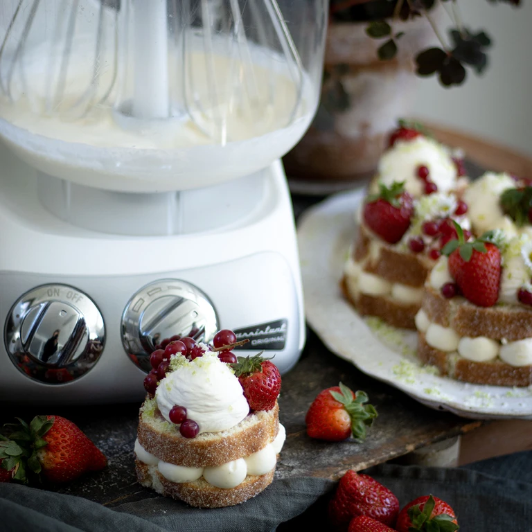 Ankarsrum Original stand mixer with whipped cream beside layered sponge cakes topped with strawberries and red currants.