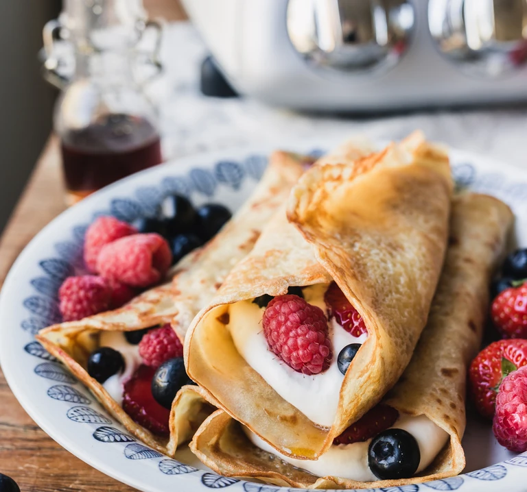Golden crepes filled with whipped cream, raspberries, blueberries, and strawberries on a decorative plate, made with Ankarsrum mixer.