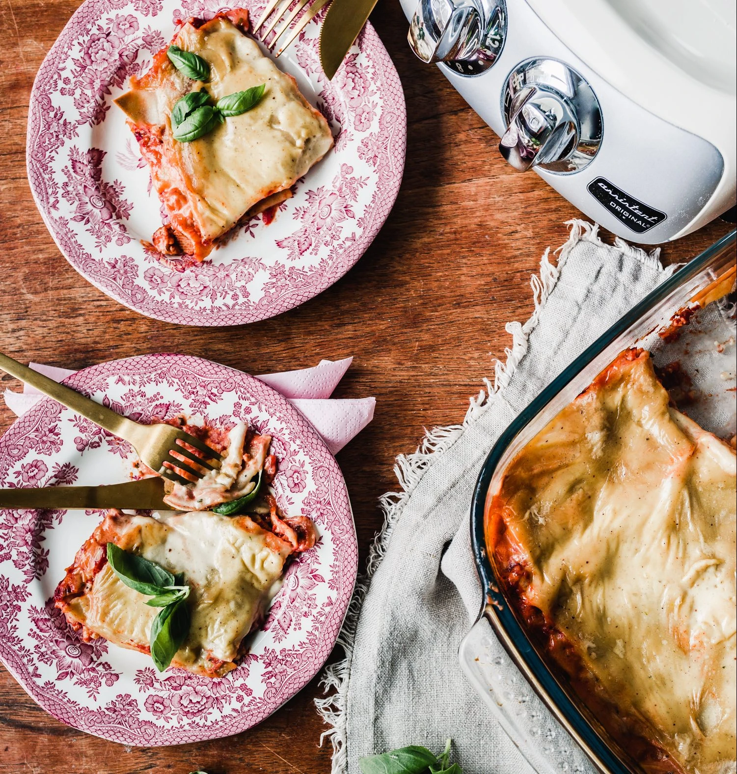 Two plates of lasagna with basil beside a baking dish and Ankarsrum Assistant Original stand mixer on a wooden table.