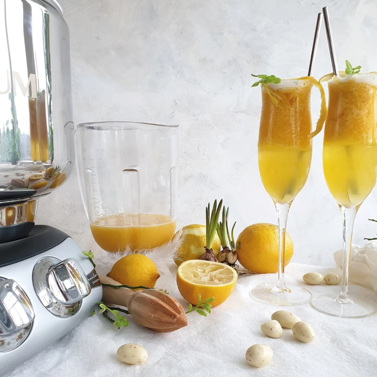 Ankarsrum blender making fresh lemon cocktails, with lemons, wooden juicer, and elegant flute glasses on white surface.