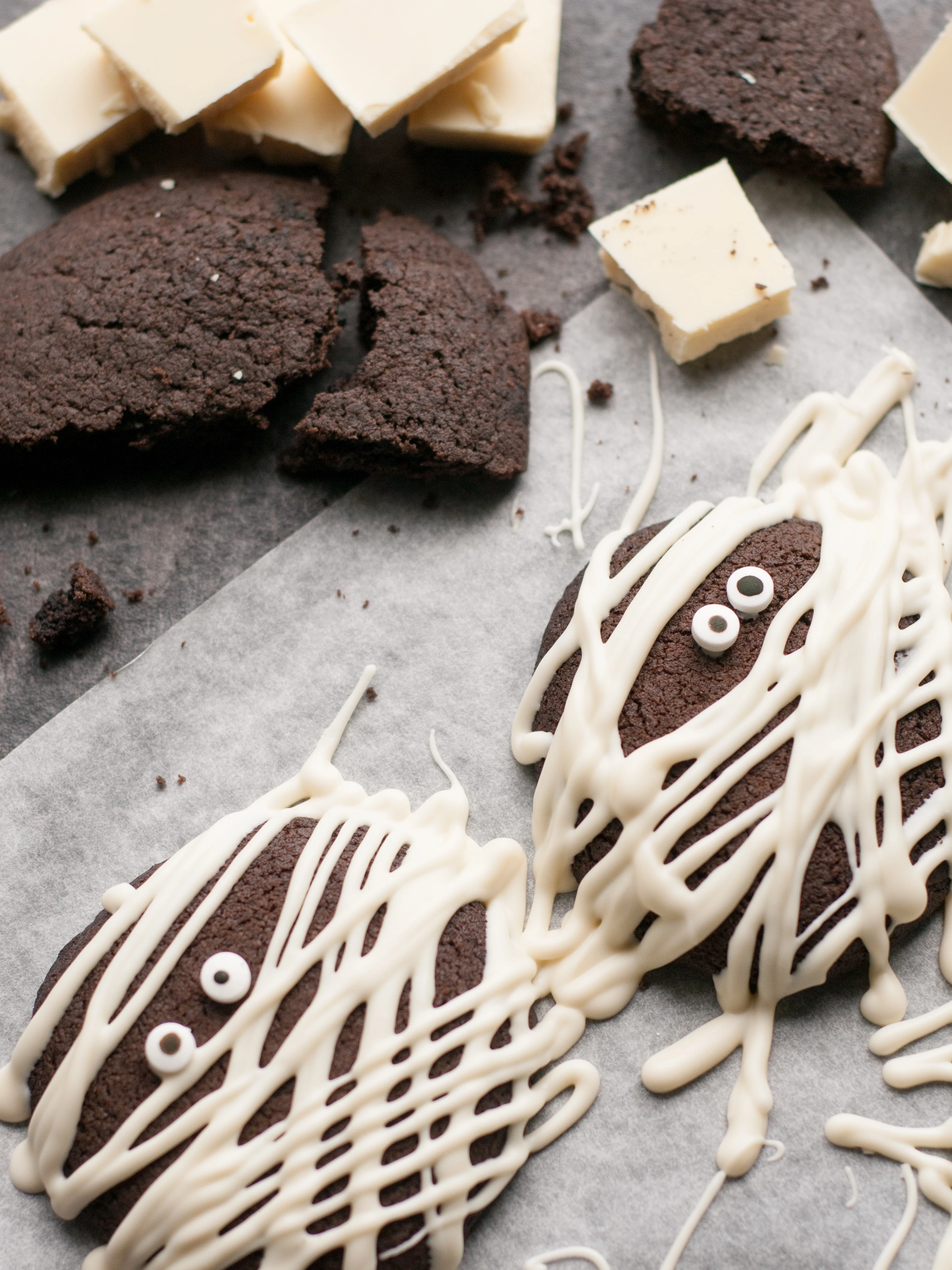 Chocolate mummy cookies drizzled with white chocolate and candy eyes, with white chocolate pieces on parchment paper.