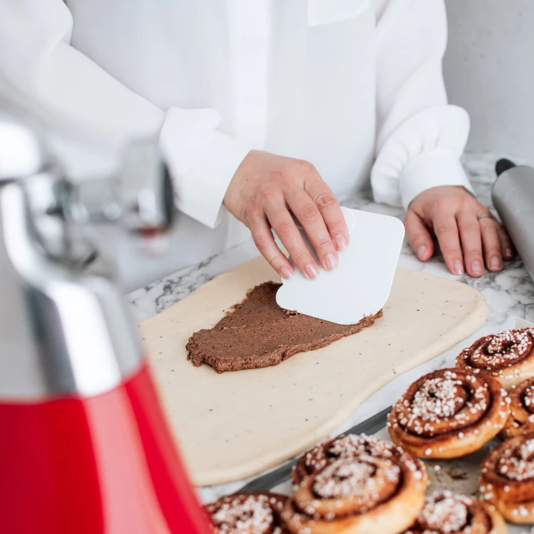 Chef spreading cinnamon roll filling on dough beside finished cinnamon rolls, using Ankarsrum stand mixer in background.