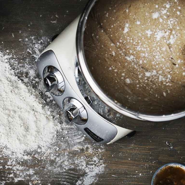 Ankarsrum stand mixer covered in flour beside a mixing bowl with bread dough on a rustic wooden surface.