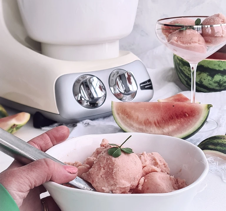 Ankarsrum stand mixer beside bowls of watermelon sorbet, fresh watermelon slices, and a coupe glass with sorbet scoops.