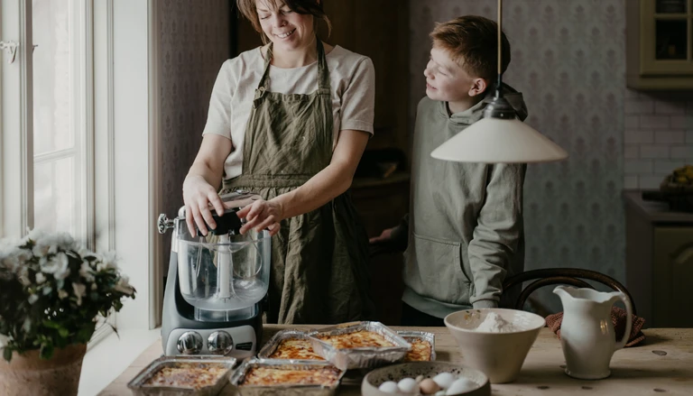 A woman and a boy cook together in a kitchen with an Ankarsrum Assistent Original. Several baked dishes and ingredients are arranged on the table, ready for cooking.