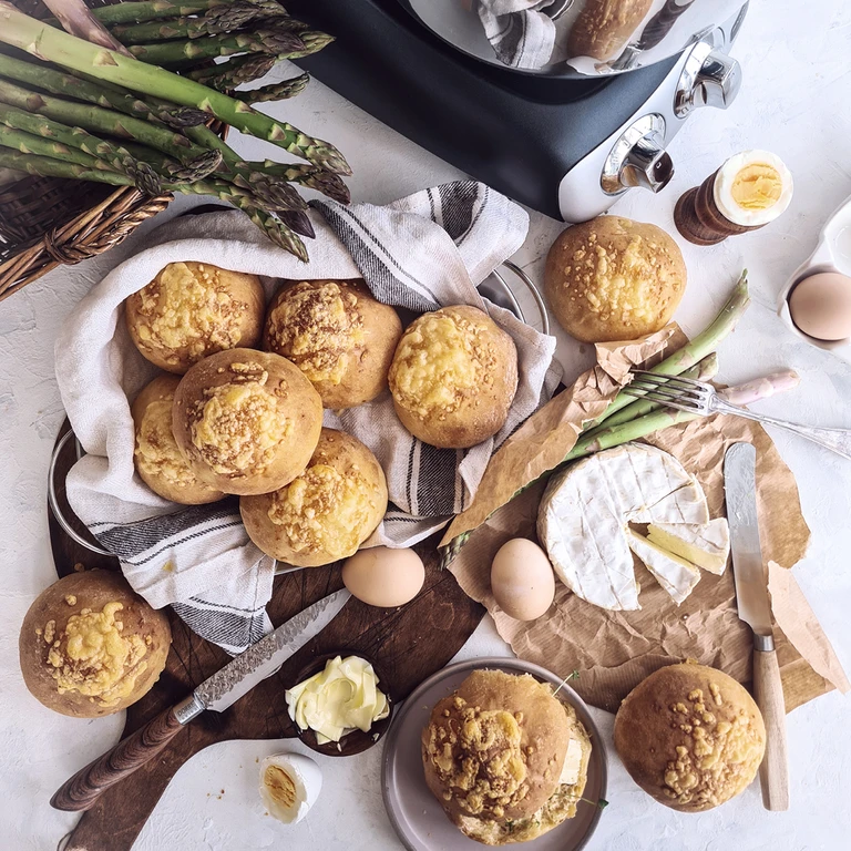 Freshly baked cheese-topped bread rolls displayed with camembert, asparagus, eggs, and butter on a rustic wooden board.