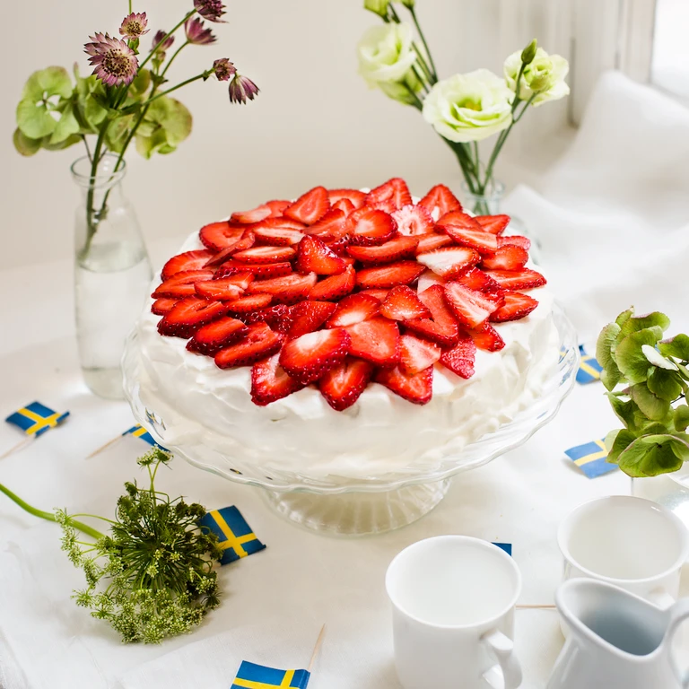 Swedish strawberry cream cake on a glass stand with Swedish flags, flowers, and white mugs — made with Ankarsrum mixer.