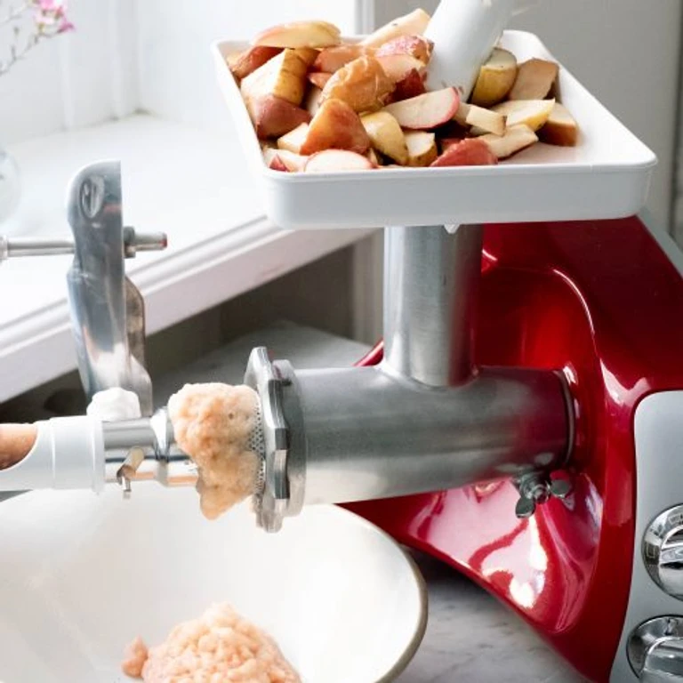 Ankarsrum red stand mixer with meat grinder attachment processing fresh apple slices into applesauce in a white bowl.