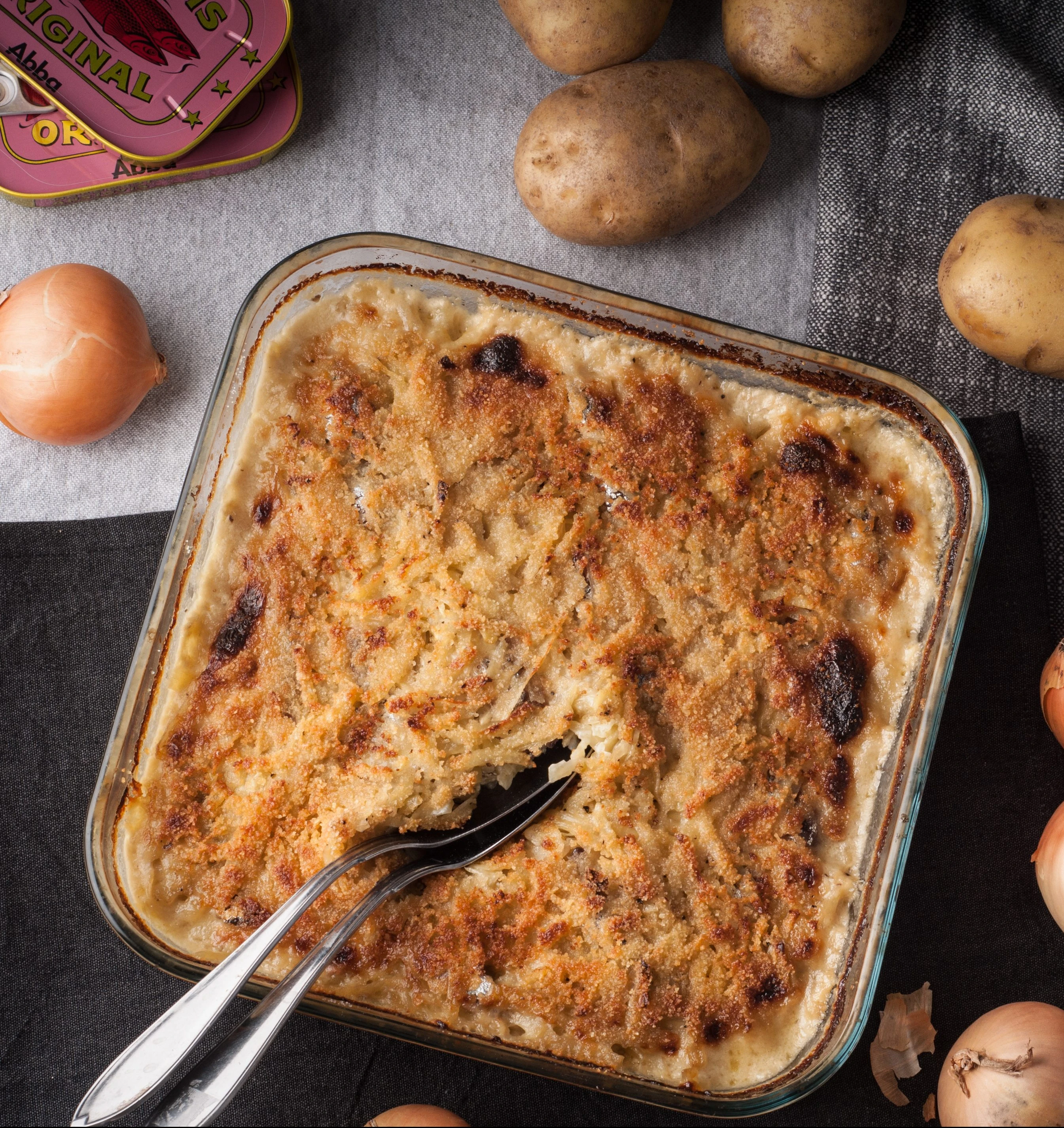 Jansson's Temptation casserole with golden crust, surrounded by potatoes, onions, and Abba anchovies tin on linen cloth.