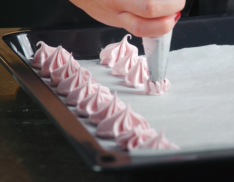 Pink meringue cookies being piped onto parchment-lined baking sheet using a pastry bag with star tip.