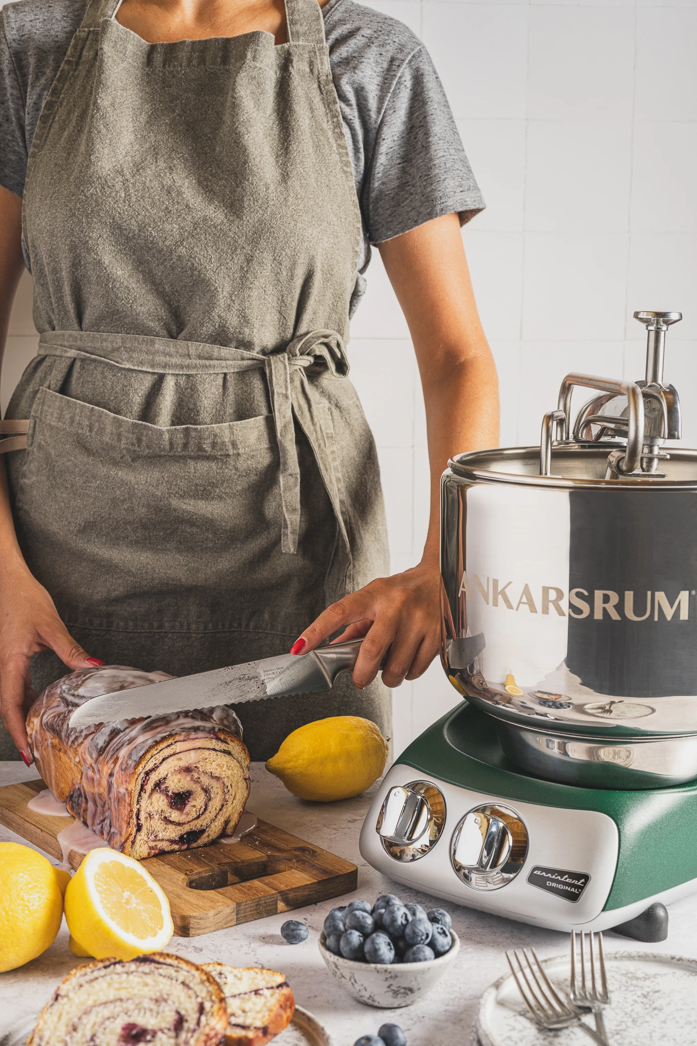 Person slicing glazed blueberry lemon swirl bread beside an Ankarsrum Original stand mixer with lemons and blueberries nearby.