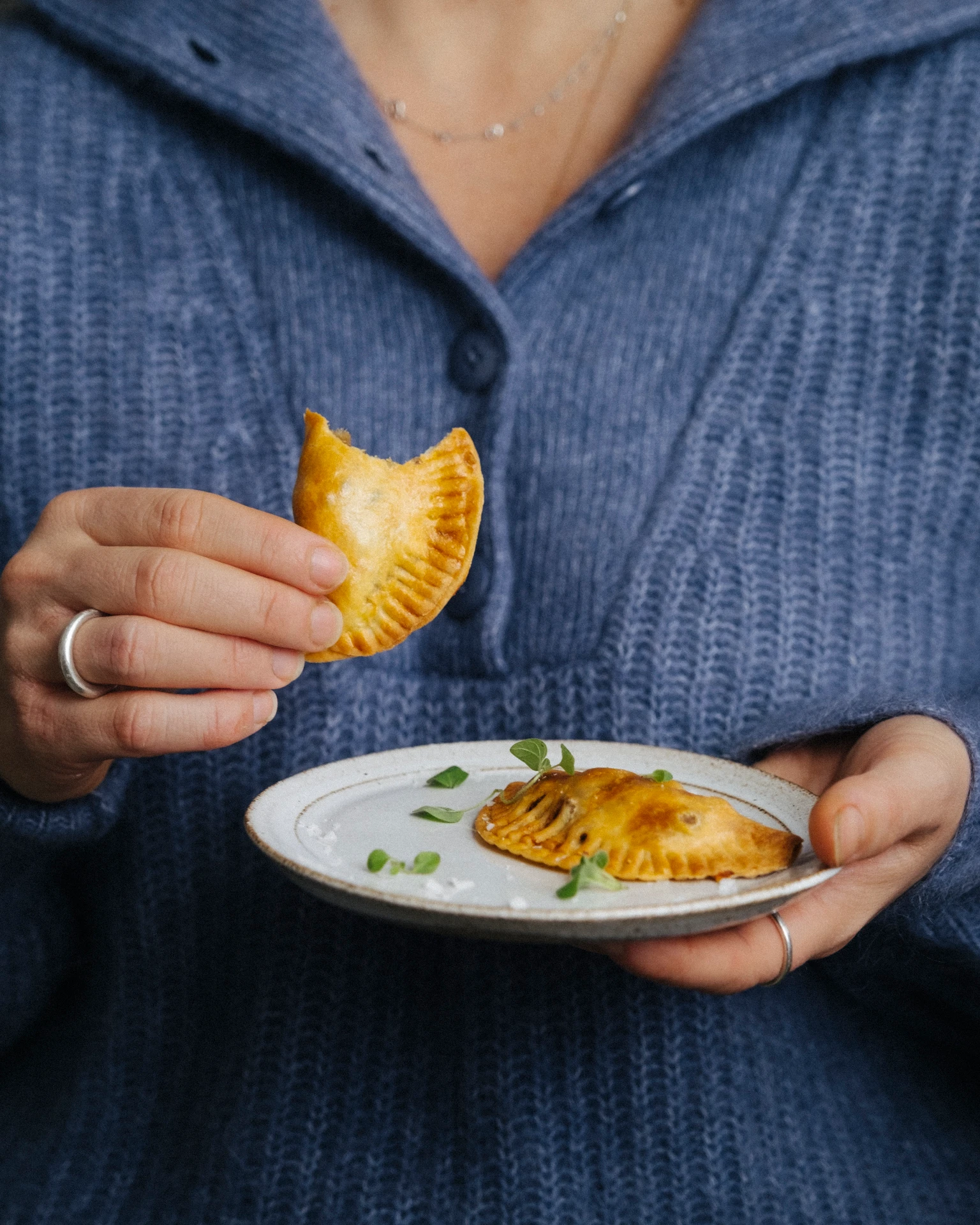 Woman in blue sweater holding a plate with golden baked empanadas, one bitten, garnished with fresh herbs.