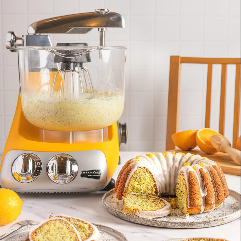 Ankarsrum Assistent Original sunbeam yellow stand mixer with lemon poppy seed bundt cake, white glaze, and fresh lemons on marble counter.