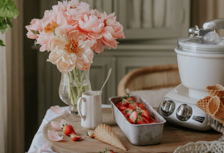Ankarsrum Original stand mixer with ice cream bowl attachment beside fresh strawberries, waffle cones, and pink peonies on a rustic wooden table.