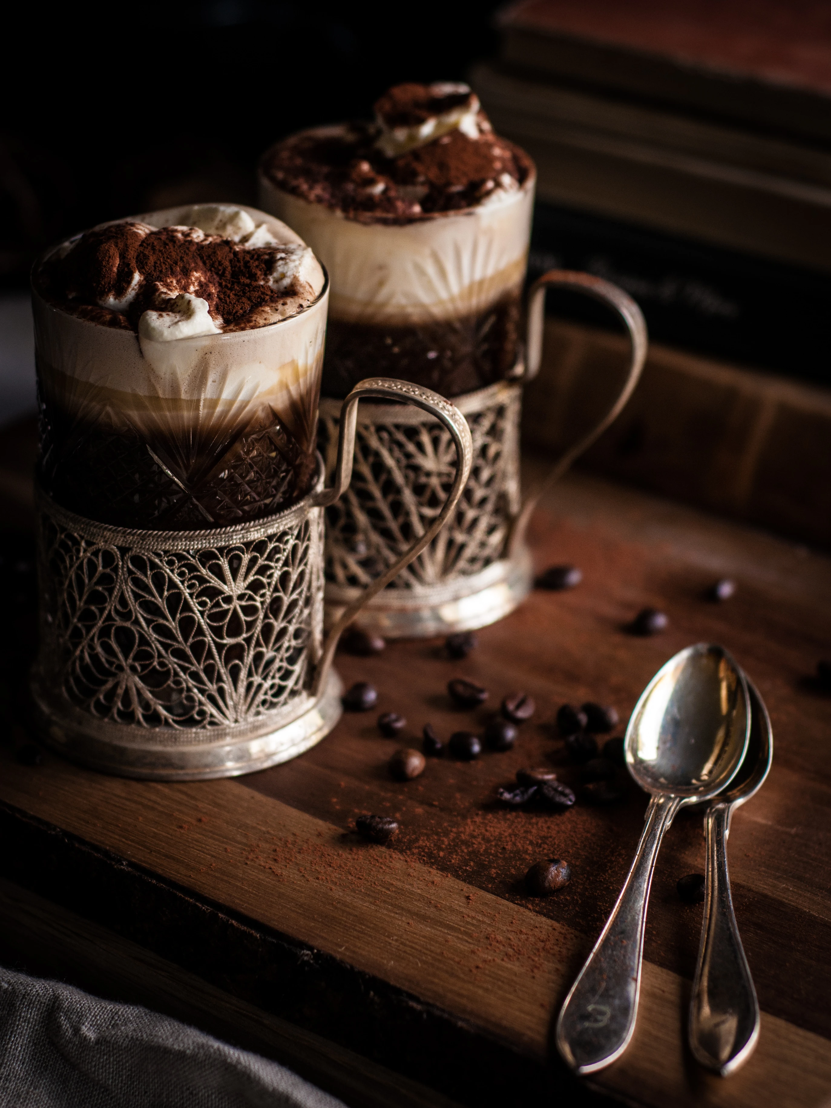 Two elegant tiramisu desserts in ornate silver glass holders on a wooden board with coffee beans and spoons.