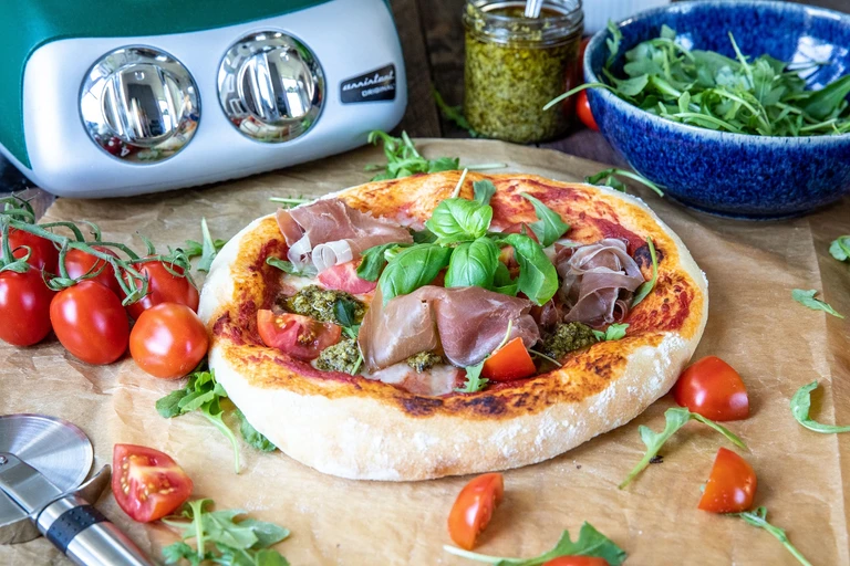 Freshly baked pizza topped with prosciutto, pesto, arugula, and tomatoes, made with an Ankarsrum Original stand mixer.