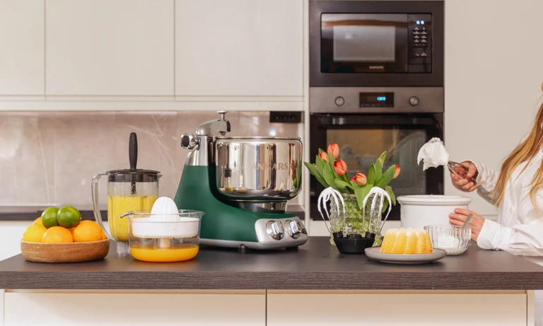 Ankarsrum stand mixer in forest green with juicer attachment, bundt cake, fresh oranges, blender attachment, and balloon whisks with whipped cream on kitchen counter.