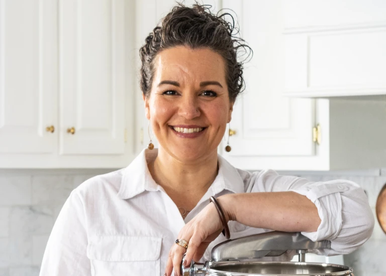 Vice president, Ashley McCord, smiling in a kitchen, leaning on an Ankarsrum stainless steel stand mixer lid, wearing a white shirt.