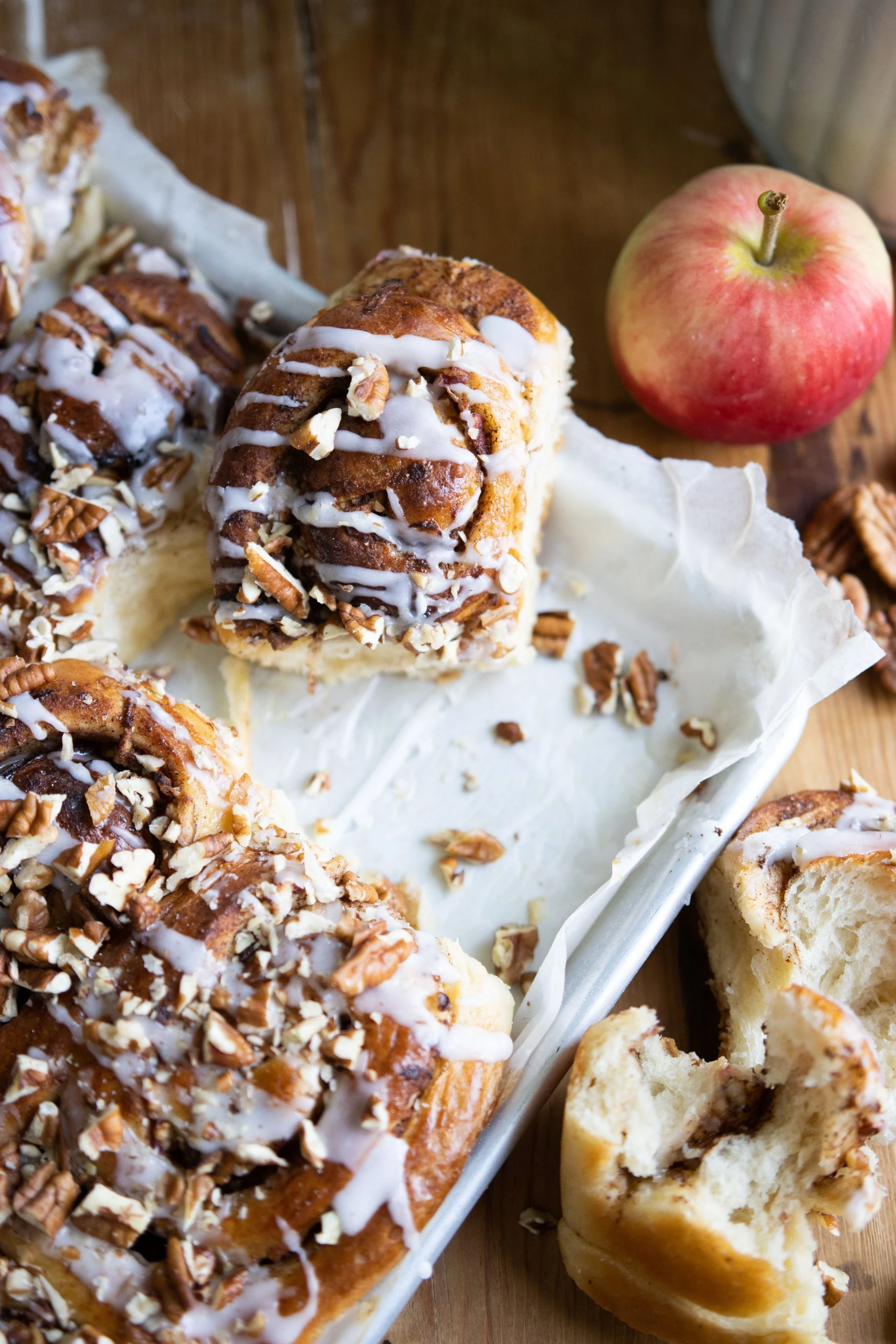 Apple pecan cinnamon rolls with white icing glaze on parchment paper, with a fresh red apple, made with Ankarsrum stand mixer.