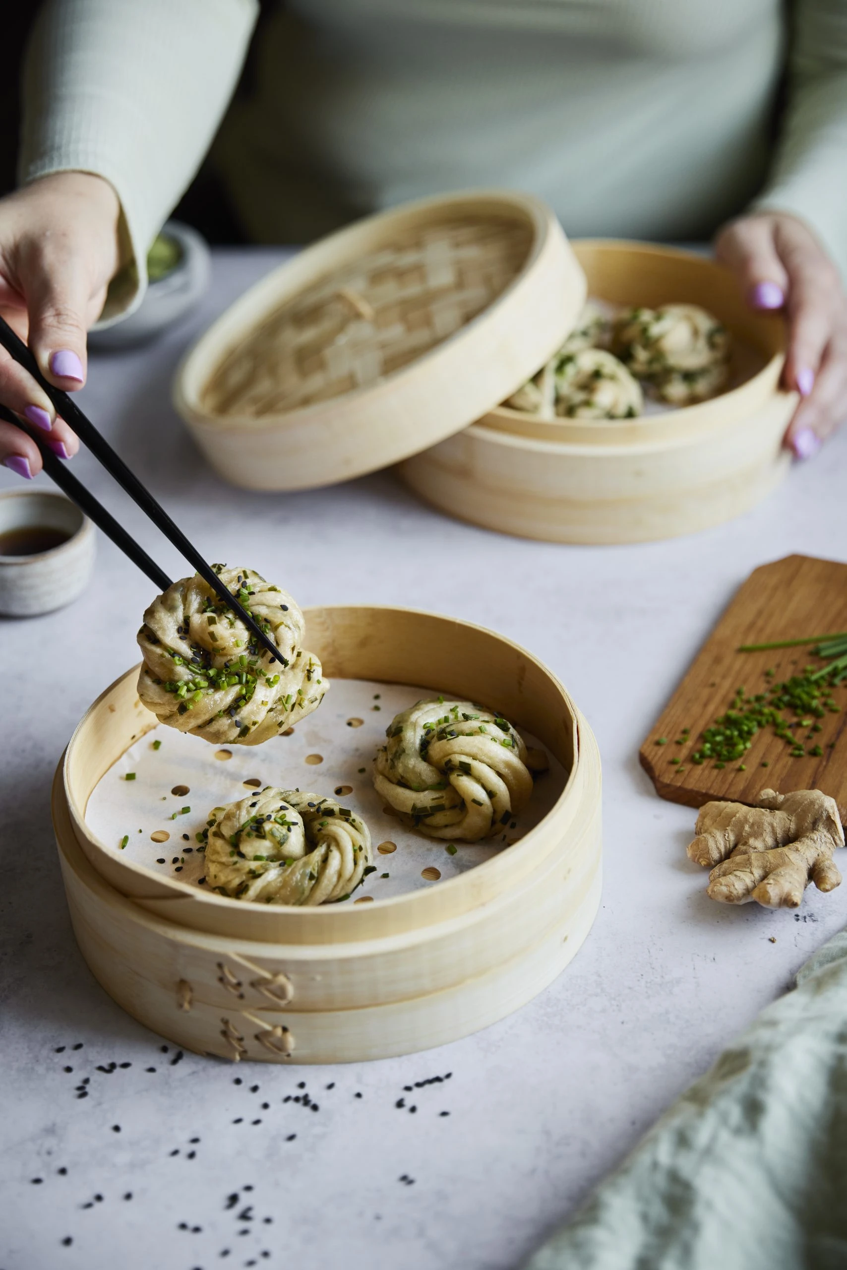 Hands using chopsticks to lift a steamed scallion bun from a bamboo steamer, with ginger and chives nearby.