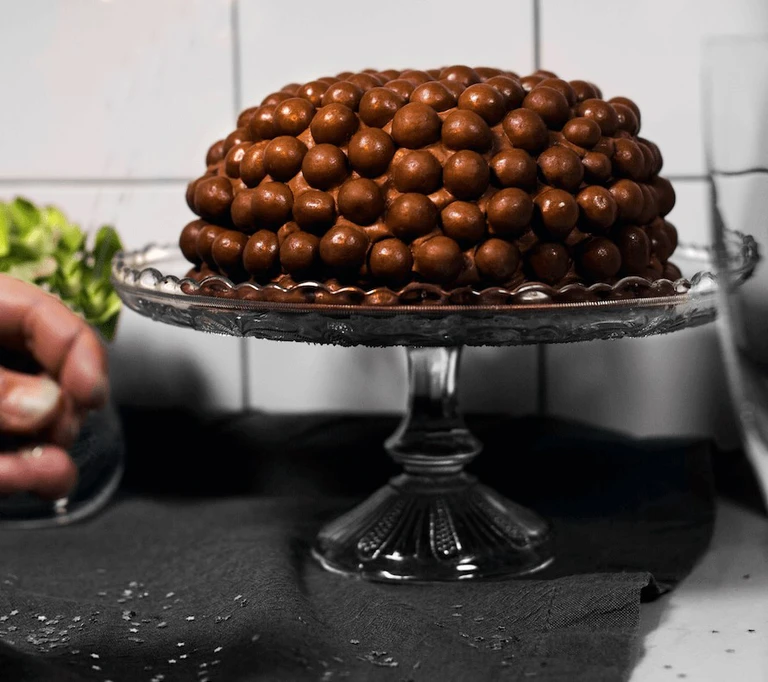 Chocolate Malteser cake covered in milk chocolate balls, displayed on an elegant glass cake stand.