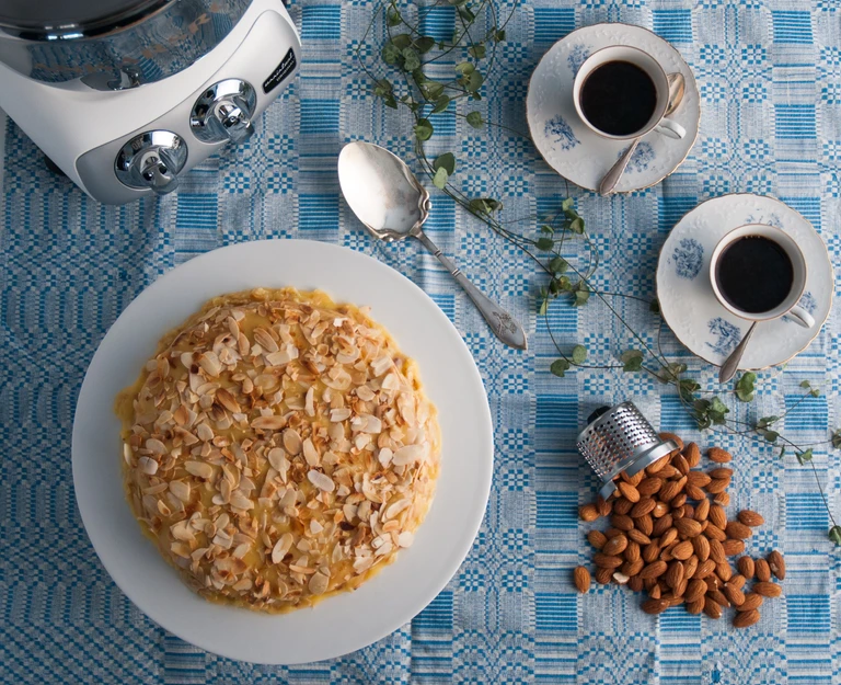 Almond-topped cake on white plate beside two espresso cups, almonds, and Ankarsrum stand mixer on blue tablecloth.