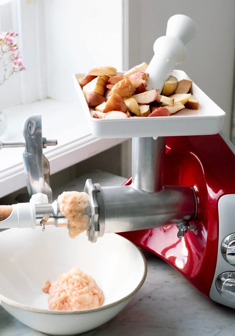 Ankarsrum stand mixer with meat grinder attachment processing fresh red apple chunks into applesauce in a white bowl.