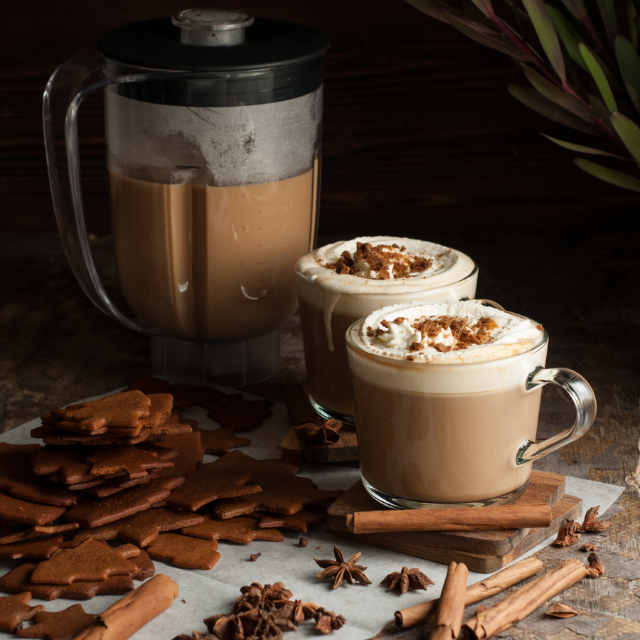 Två glas kryddig latte toppad med vispgrädde och krossade pepparkakor, serverade med pepparkakor och kanelstänger bredvid en mixerkanna.