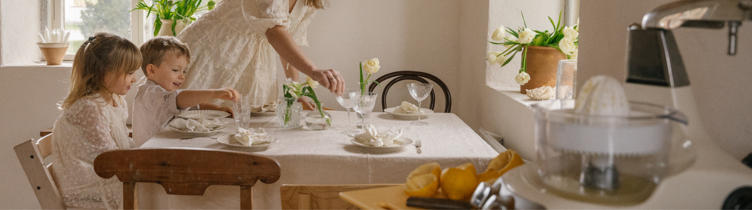 Två barn dukar ett festligt bord med vita tulpaner och linneservetter i ett ljust kök, med en Ankarsrum Assistent Original och citroner i förgrunden – en varm stund inför vårens högtider.