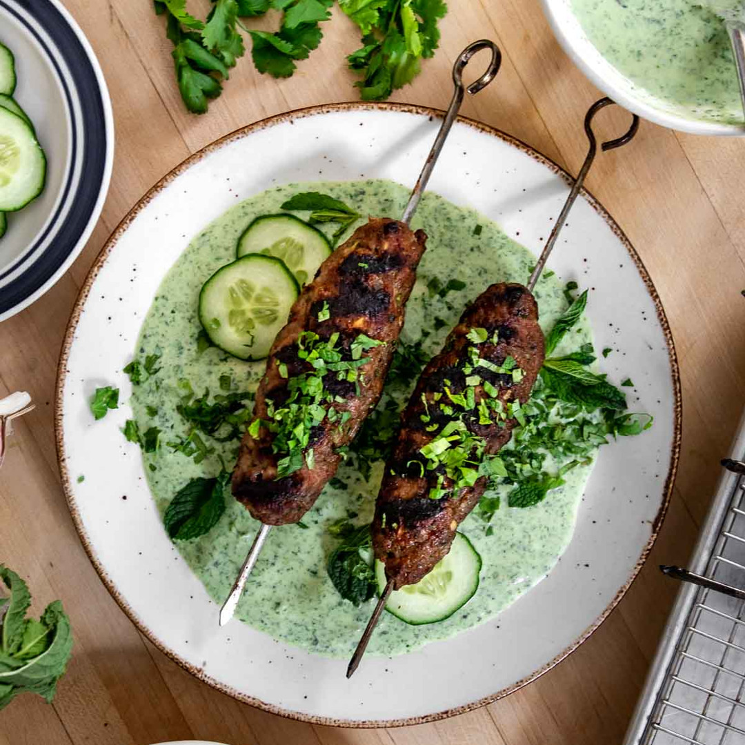 Zwei gegrillte Kebabspieße, serviert mit einer würzigen Joghurtsauce, Gurke und frischen Kräutern auf einer weißen Platte.