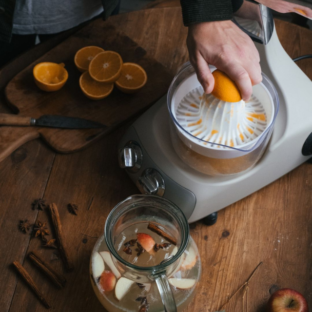 Ankarsrum kรถksmaskin med citruspress som pressar fรคrsk apelsinjuice, bredvid en karaff med รคppel- och krydddryck.