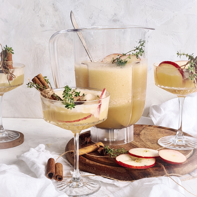 Karaff och glas med รคppelmustdrink dekorerad med kanelstรคnger, รคppelskivor och fรคrsk timjan.