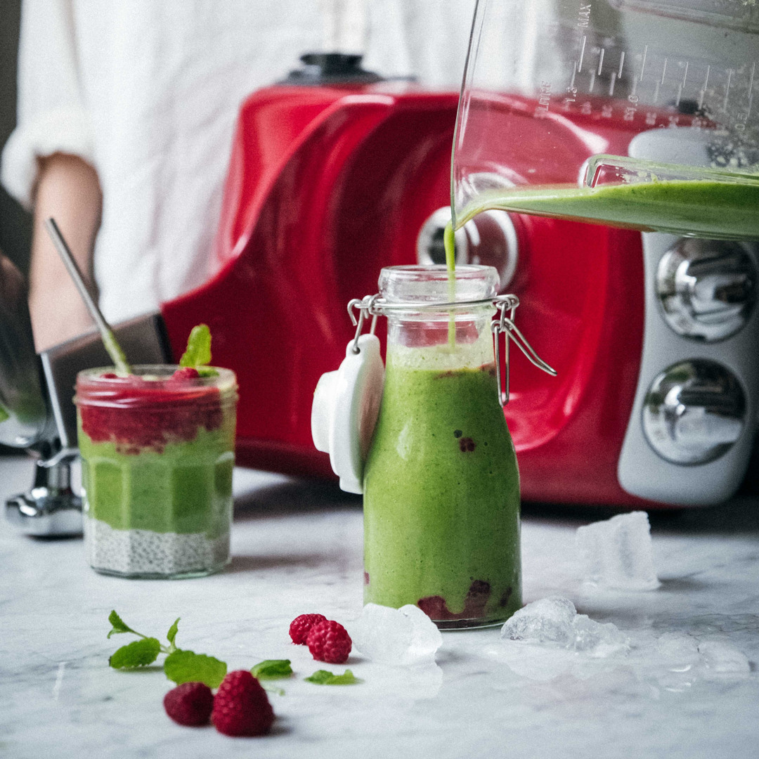 En grön smoothie hälls upp i en glasflaska framför en Ankarsrum Assistent Original, bredvid en chiapudding toppad med hallon.