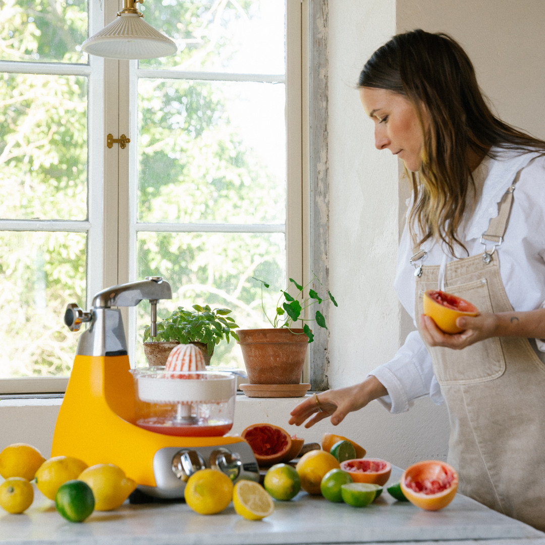 En kvinna använder Ankarsrums Citruspress monterad på en Ankarsrum Assistent Original i Sunbeam Yellow för att pressa färsk citrusjuice. Citroner, lime och grapefrukt ligger på köksbänken.
