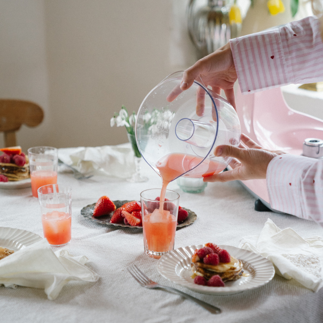 Person häller upp en rosa jordgubbsdryck i ett glas vid ett dukat bord med färska bär, pannkakor och flera glas dryck. En Ankarsrum Assistent Original syns i bakgrunden.