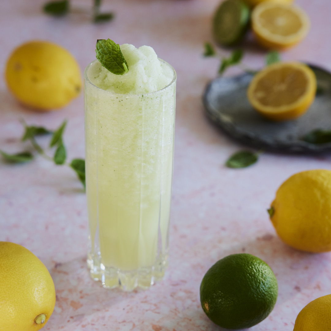 Högt glas med ljusgul citrusslush toppad med färskt myntablad. Citroner, lime och mynta ligger utspridda runt glaset på en ljus yta.