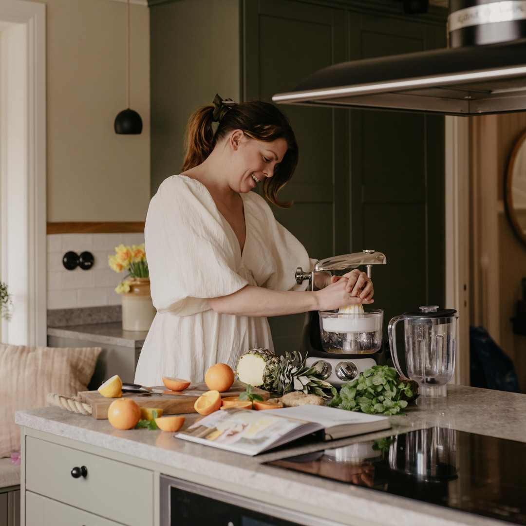 Kvinna i vit klänning pressar citrusfrukter med en Ankarsrum Assistent original på en köksö. Färska apelsiner, ananas, ingefära och örter ligger framme bredvid en öppen receptbok i ett modernt kök.