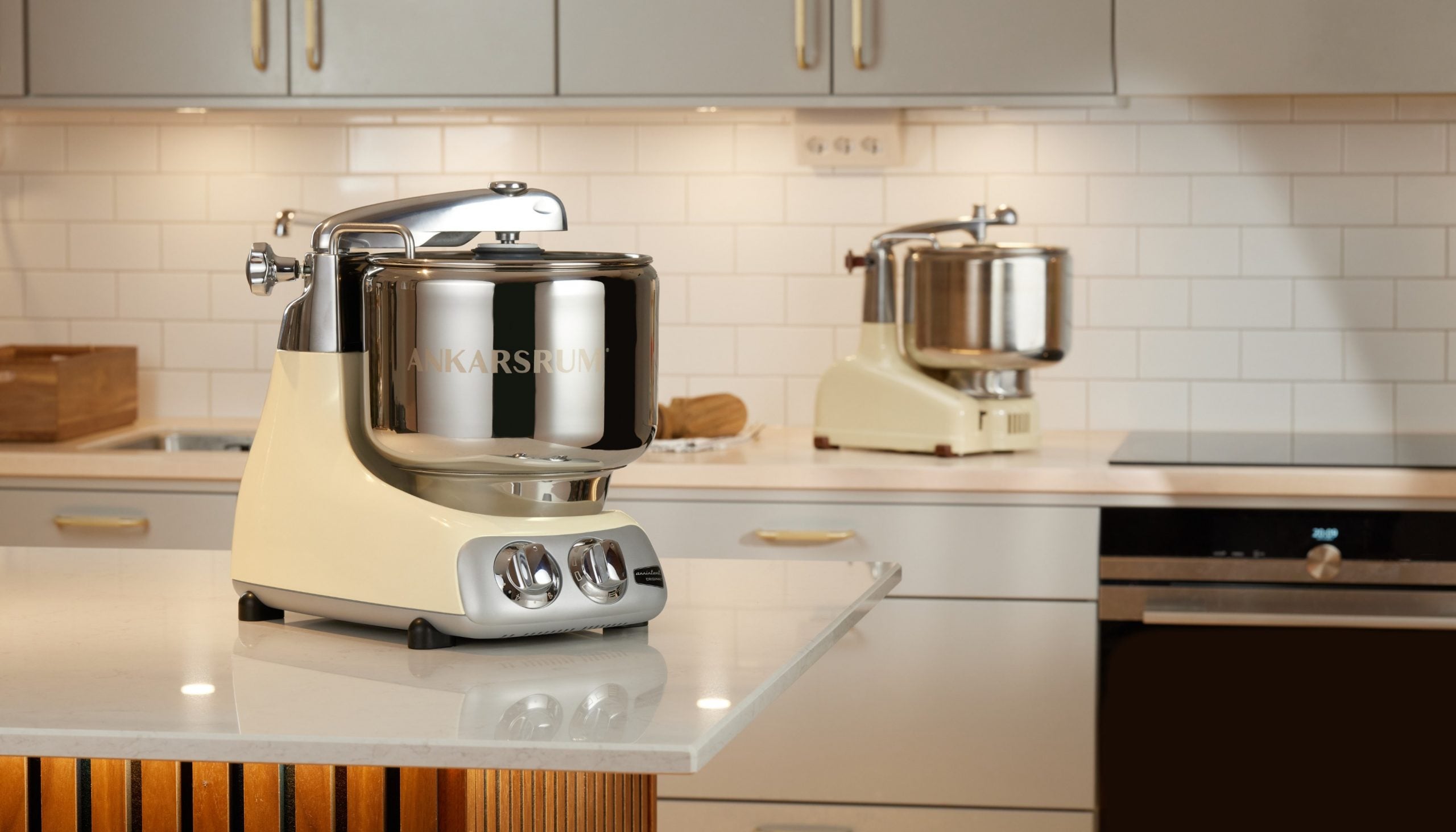 Ankarsrum stand mixer in cream with chrome bowl on white kitchen countertop, second mixer visible in background.