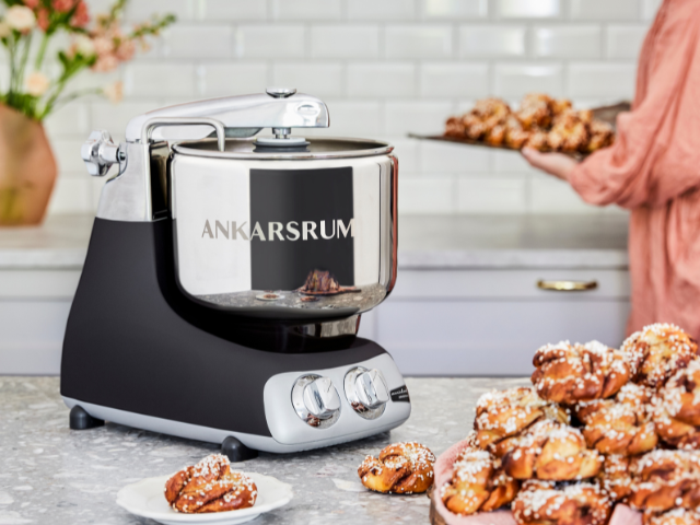 Ankarsrum stand mixer in the colour Black with stainless steel bowl, placed on a kitchen counter next to freshly baked buns