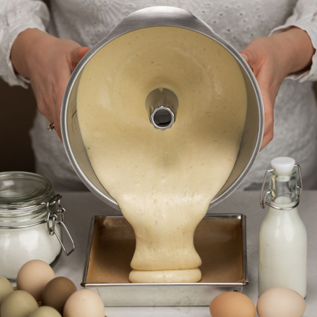 Cake batter being poured from an Ankarsrum Beater Bowl in Stainless Steel into a rectangular baking pan. Eggs, sugar, and milk are arranged on the countertop around the pan. Durable stainless steel bowl designed for stable whipping, precision mixing, and reliable performance in baking.