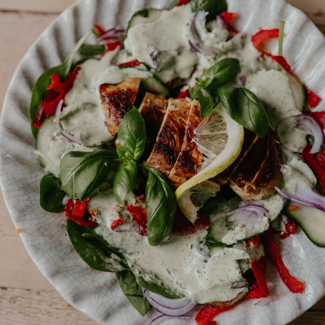 Plate of chicken salad with spinach, cucumber, bell pepper, and red onion. The salad is topped with basil, lemon, and creamy herb dressing.