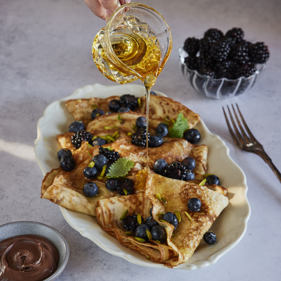 Honey is poured over folded crêpes topped with blueberries, blackberries, and pistachios on a serving platter. A bowl of blackberries and a bowl of chocolate cream sit alongside.