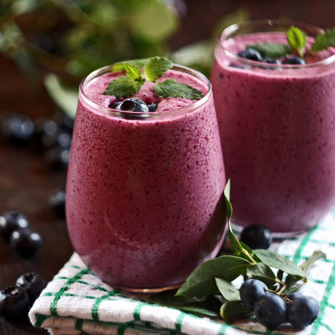 Two glasses of purple blueberry smoothie garnished with fresh blueberries and mint leaves. Blueberries and green leaves are arranged around the glasses on a table with a kitchen towel in the foreground.
